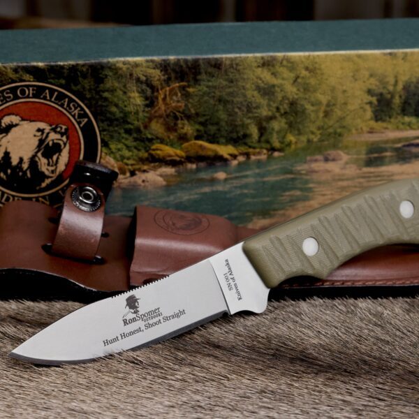 Ron Spomer edition Classic Hunter drop point hunting knife with green textured handle, engraved blade, leather sheath, and Knives of Alaska branded box on a natural fur surface.