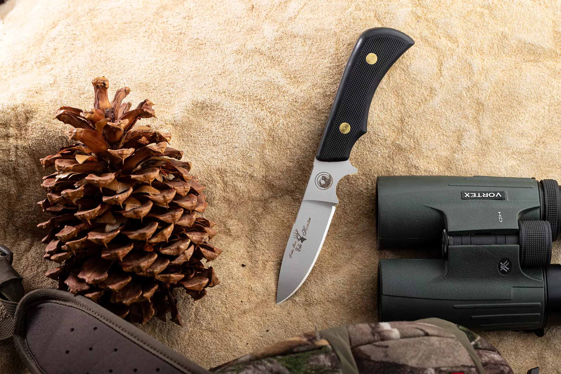 Trekker Elk Hunter knife with Suregrip handle displayed on tan fabric beside a pine cone and Vortex binoculars