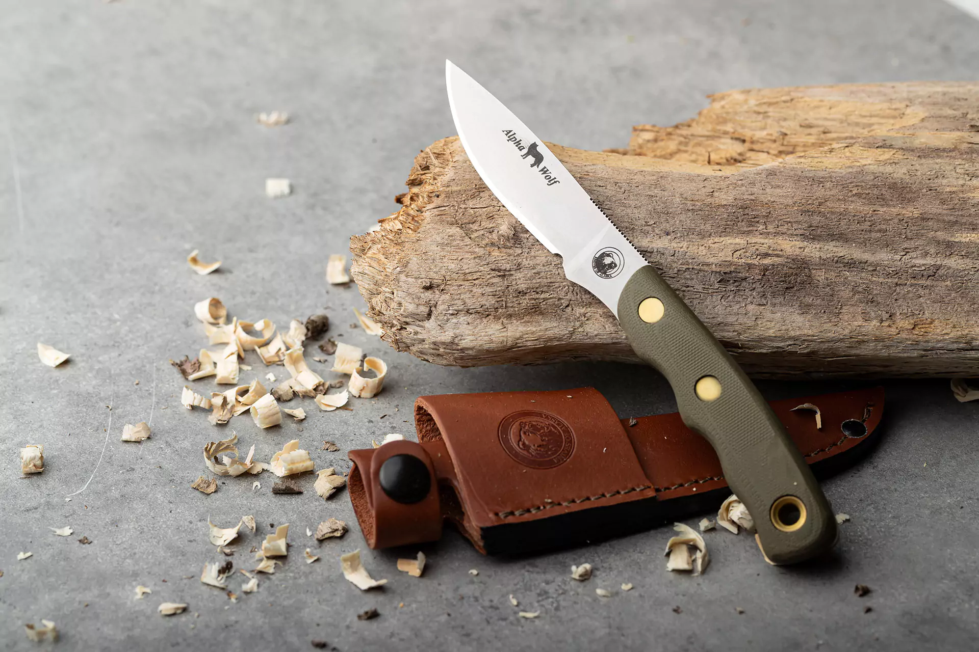 Alpha Wolf knife with OD green G10 handle resting on wood beside leather sheath and wood shavings
