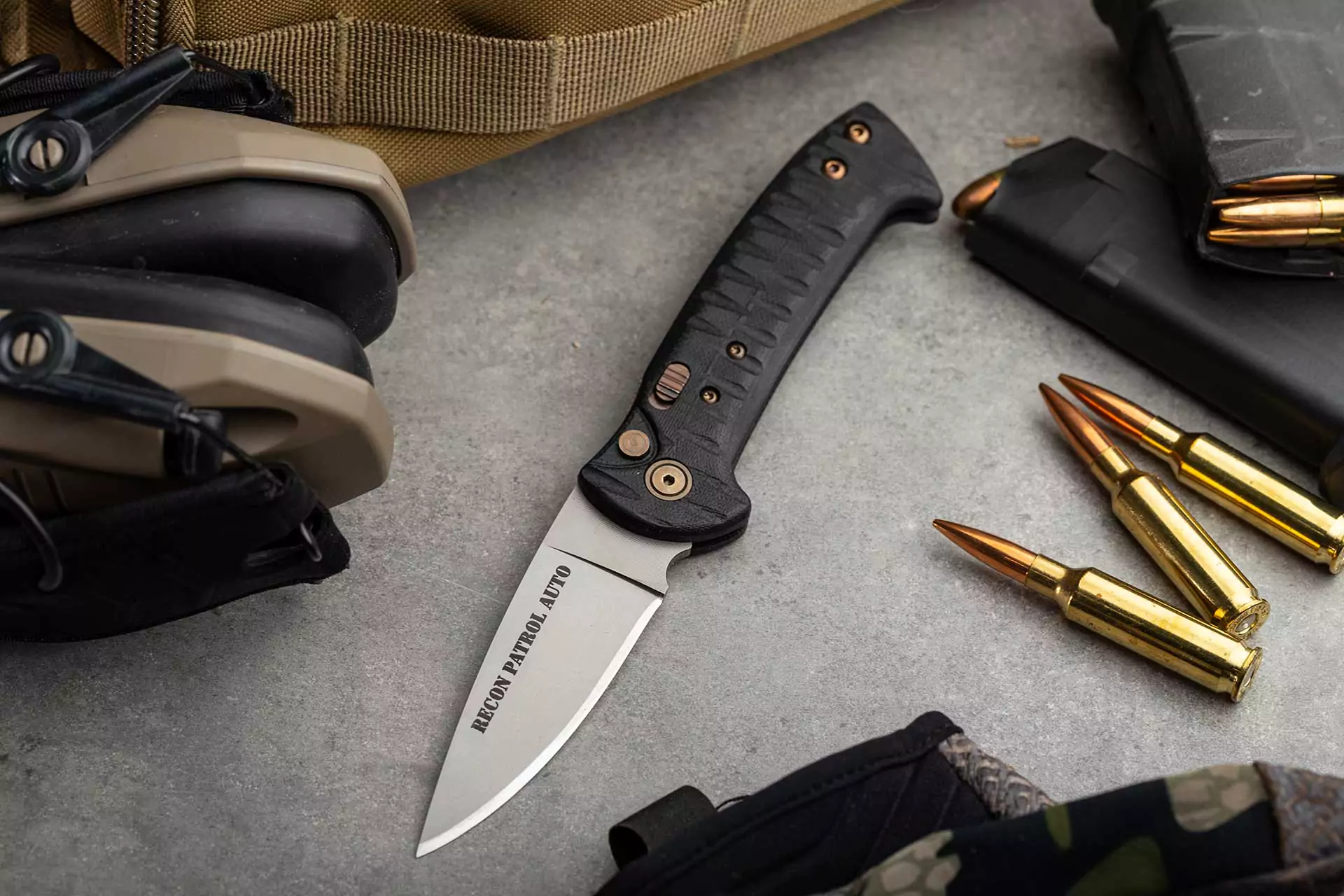Recon Patrol Auto knife with Magnacut steel blade and G10 handle beside rifle rounds and tactical gear
