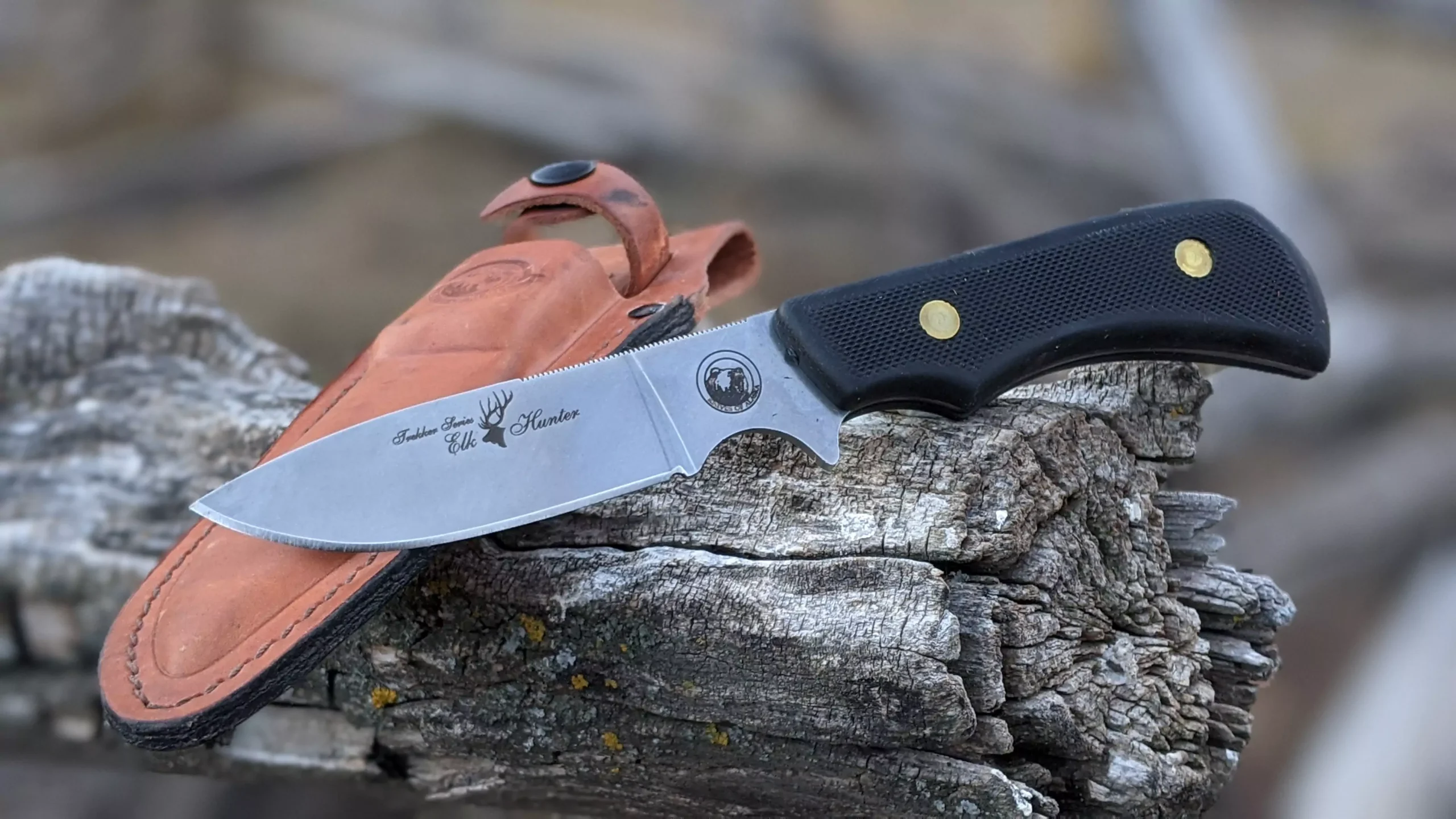 Trekker Elk Hunter knife with black SureGrip handle resting on weathered wood with leather sheath